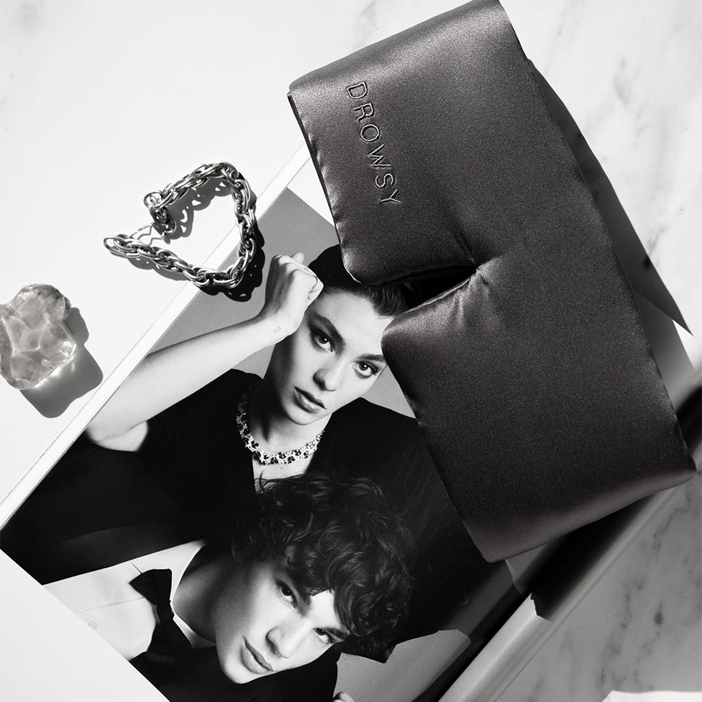 The Moonlight Shadow silk sleep mask by drowsysleepco, embroidered with “DROWSY,” rests on a black-and-white fashion magazine, beside a silver chain bracelet and translucent stone on marble—promising truly beautiful sleep.