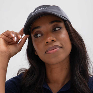 A woman with long dark hair, calm and thoughtful, wears the Drowsy Silk Cap - Moonlight Shadow by Drowsy. She touches the brim of the stylish dark sun shield cap, her hair protected and face shaded.