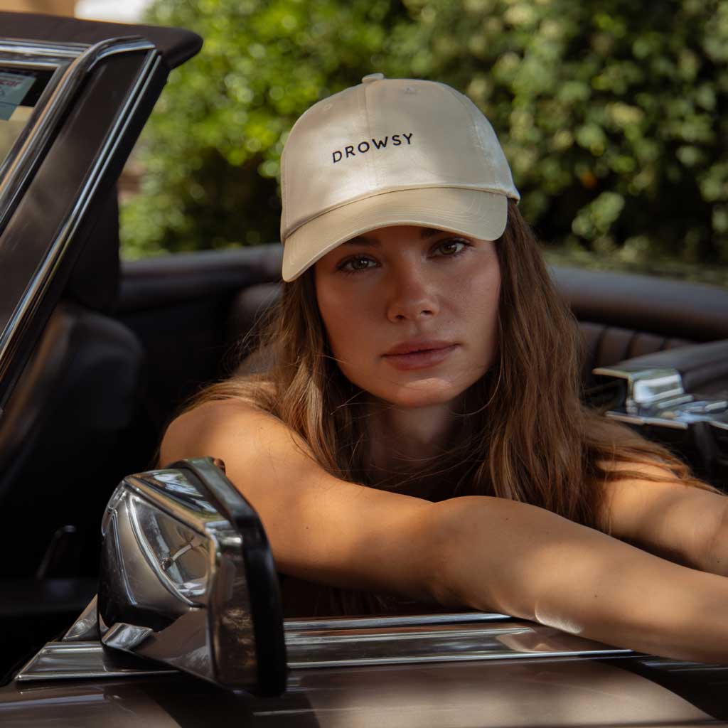 A woman with long hair wears the drowsysleepco Perfect Escape Bundle in Dusty Gold and sits in a convertible, exuding post-beauty sleep confidence as she looks at the camera, green foliage in the background.