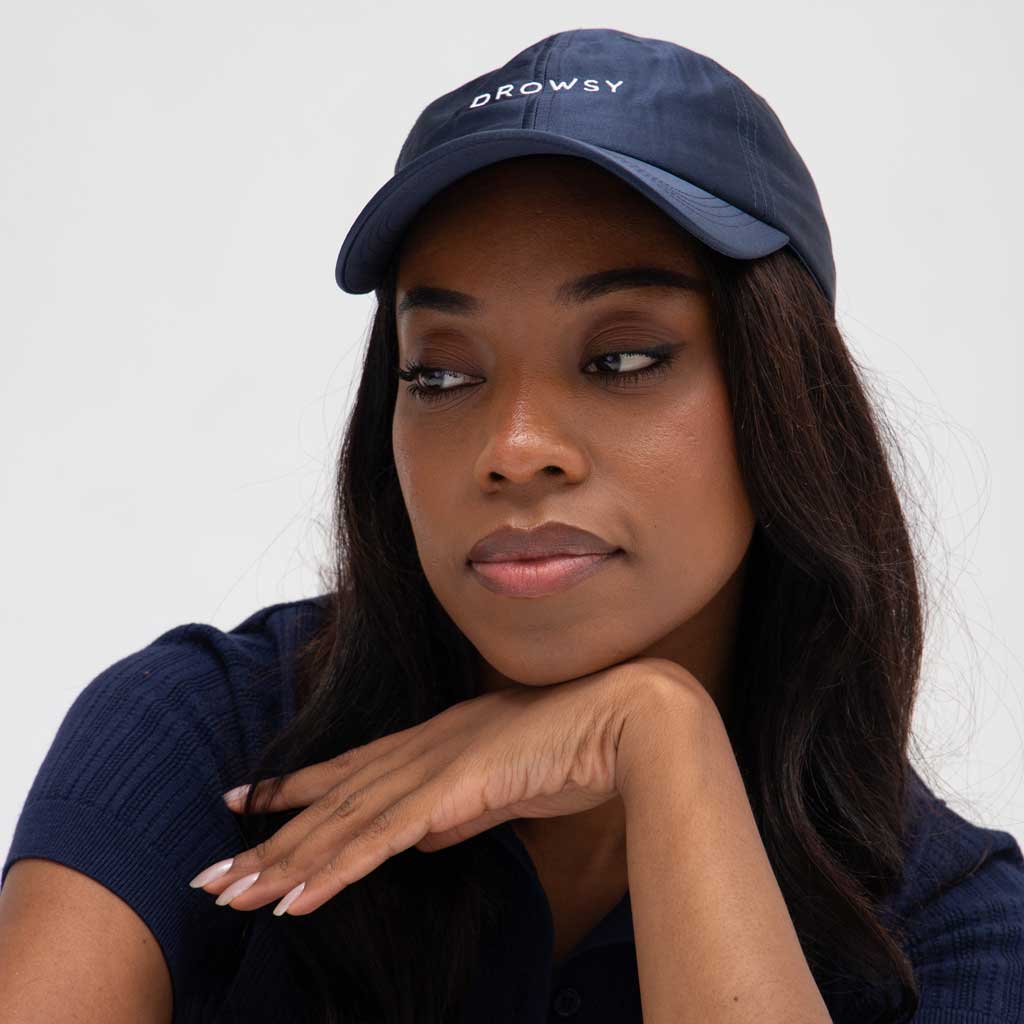 A woman with long dark hair wears the Drowsy Silk Cap in Midnight Blue and a matching polo. The cap, perfect for sunny days, features the Drowsy logo. She rests her chin on her hand, gazing thoughtfully to the side against a light gray background.