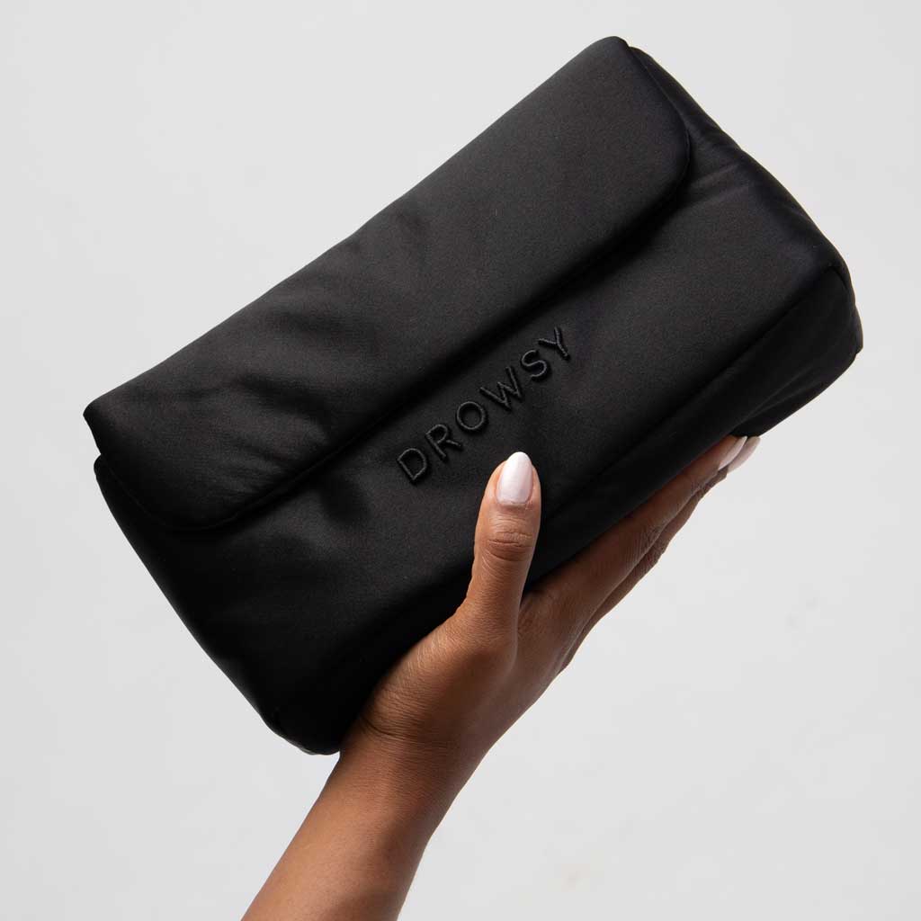 A hand holds the Drowsy Silk Necessaire - Black Jade by Drowsy, a sleek black silk pouch embossed with DROWSY on the front—perfect for elevating your beauty routine—set against a plain light grey background.