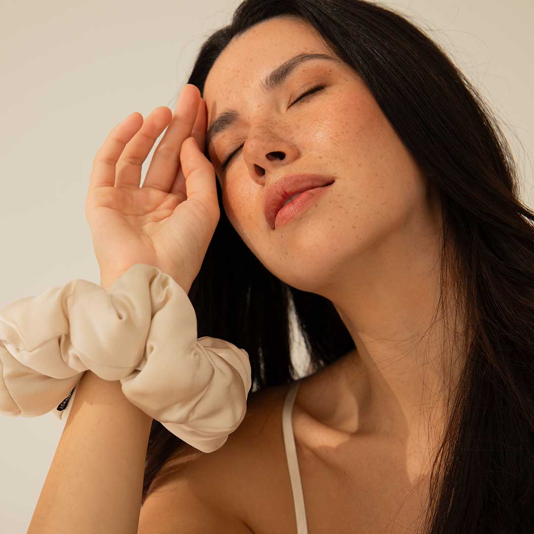 A woman with long dark hair and closed eyes touches her forehead gently with her hand, wearing a light-colored scrunchie on her wrist and a white strap top, in soft natural lighting.