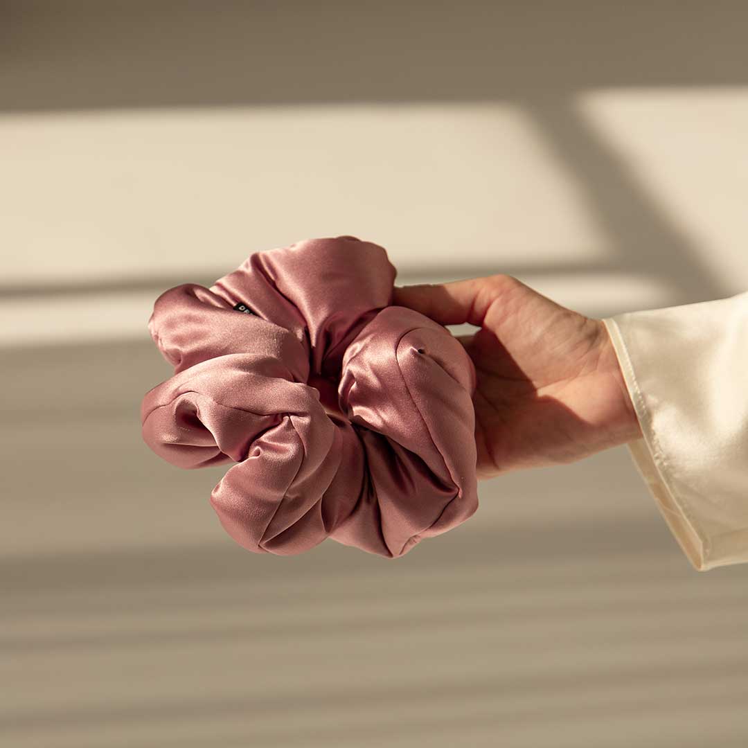 A hand in a white sleeve holds the drowsysleepco Damask Rose - Padded Scrunchie, a shiny pink silk hair accessory, against a neutral background with soft light and shadows.