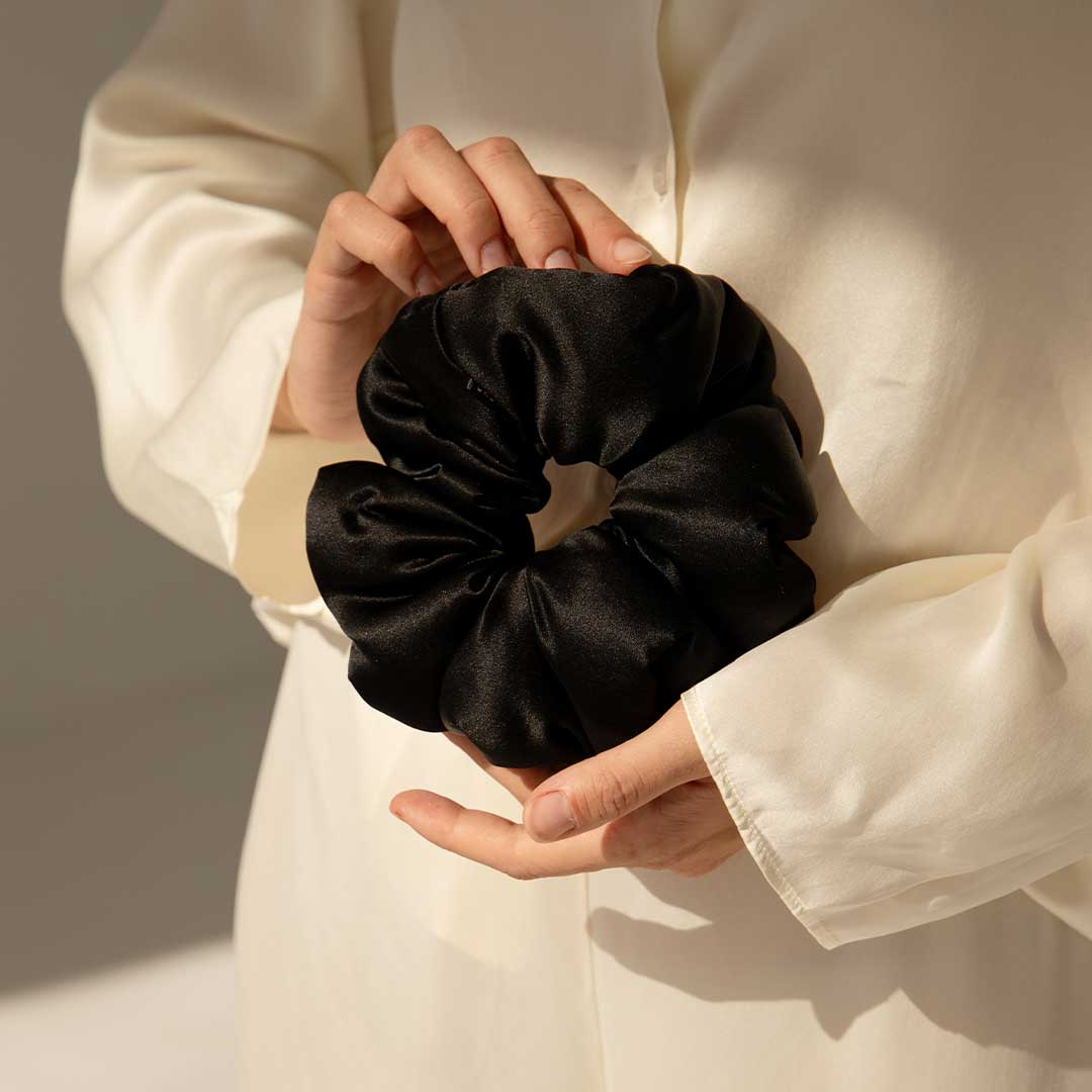 A person wearing a cream-colored shirt holds a large black satin scrunchie with both hands, against a softly lit neutral background.