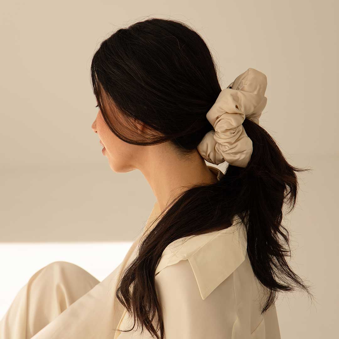 A woman with long dark hair tied in a low ponytail with the Rajah Padded Scrunchie by drowsysleepco sits in profile, wearing a cream blouse against a neutral background.