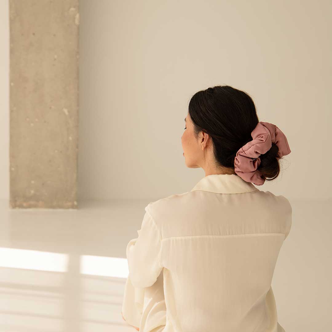 A woman with dark hair tied in a large pink scrunchie sits on the floor, facing away, wearing a cream-colored blouse in a softly lit minimalist room with neutral walls and a concrete pillar.