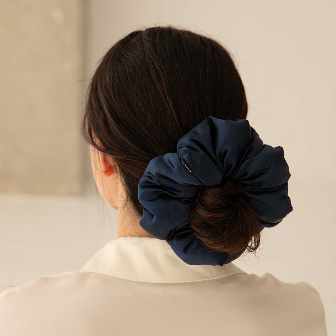 A woman with dark hair in a low bun, secured with the drowsysleepco Midnight Blue - Padded Scrunchie, is seen from behind. She wears an off-white collared shirt against a neutral background.
