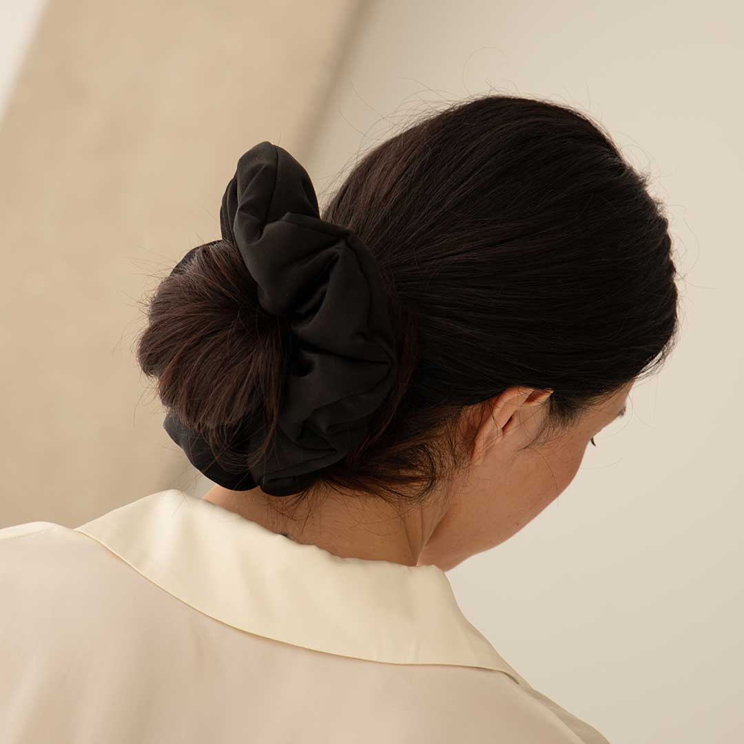 A woman with dark hair in a low bun, secured by the drowsysleepco Black Jade - Padded Scrunchie, wears a cream-colored collared shirt and is shown from behind against a neutral background.