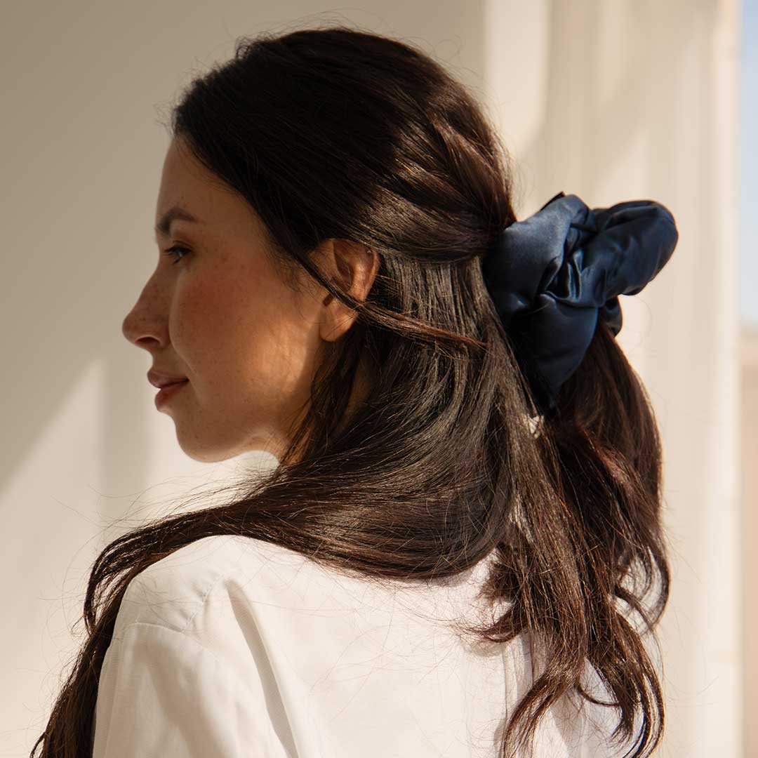 A woman with long dark hair tied in a navy blue scrunchie looks to the side, wearing a white shirt and standing in soft natural light.