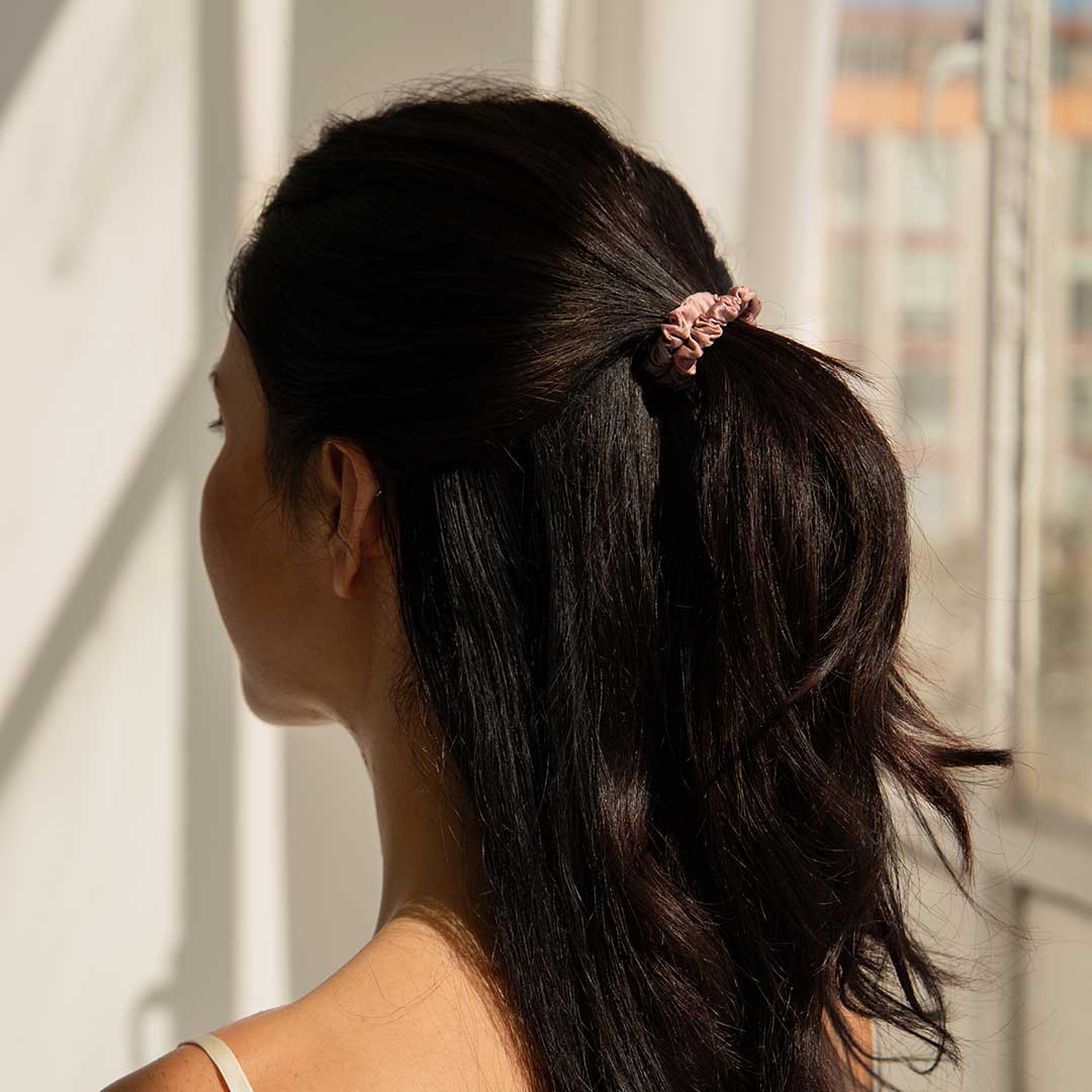 A woman with long, dark hair tied in a pink scrunchie faces away from the camera, standing near a sunlit window with sheer curtains.
