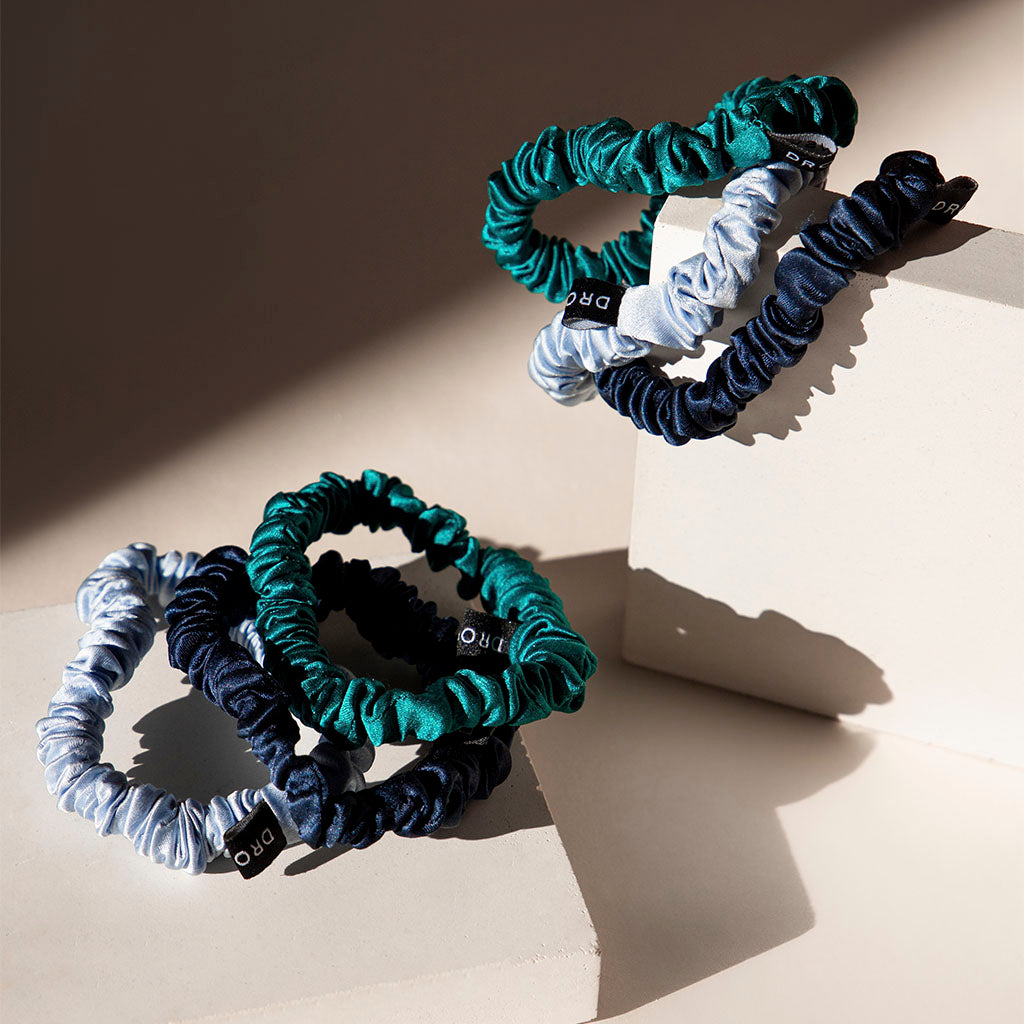 The drowsysleepco Skinny Scrunchie Set - Blue & Green Shades features three silk scrunchies in green, light blue, and navy with black tags—designed for gentle hold and hair health. Perfectly arranged on beige blocks casting soft shadows.