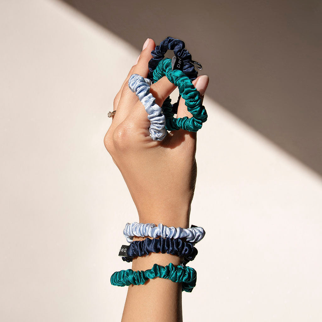 A hand displays drowsysleepcos Skinny Scrunchie Set in blue and green shades, looped around the fingers and wrist against a beige background—an elegant set designed for hair health.