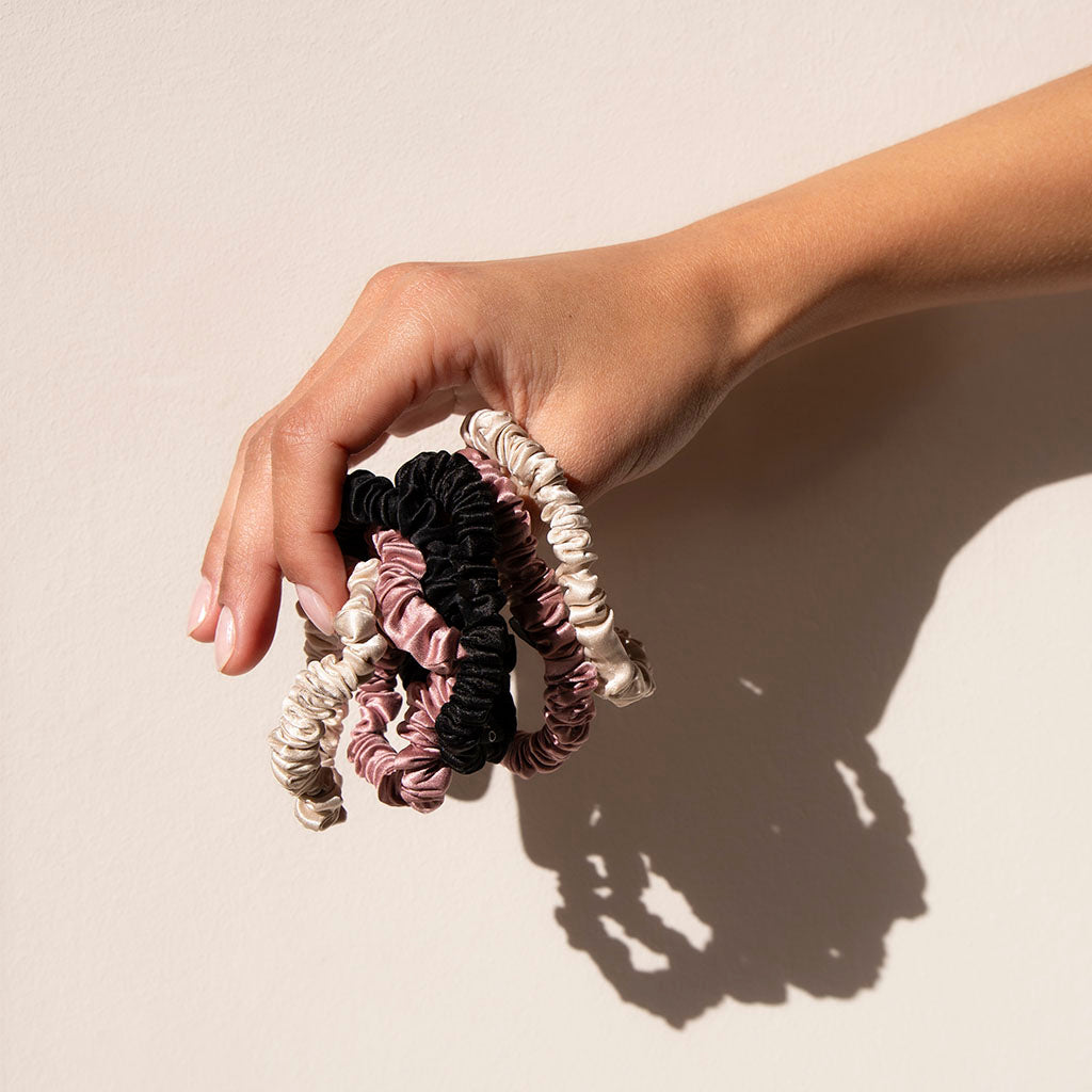 A hand displays drowsysleepcos Skinny Scrunchie Set in Rose, Dusty, and Black against a light backdrop. These 22 momme silk scrunchies are gentle on hair, promoting better hair health.