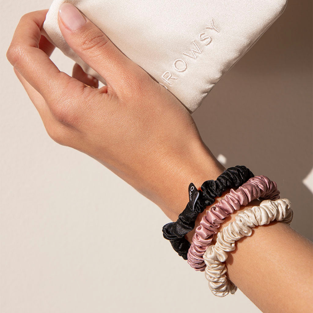 A hand wearing three drowsysleepco Skinny Scrunchies in Rose, Dusty, and Black—crafted from 22 momme silk for hair health—holds a white pouch with ROWSY embroidered on it.