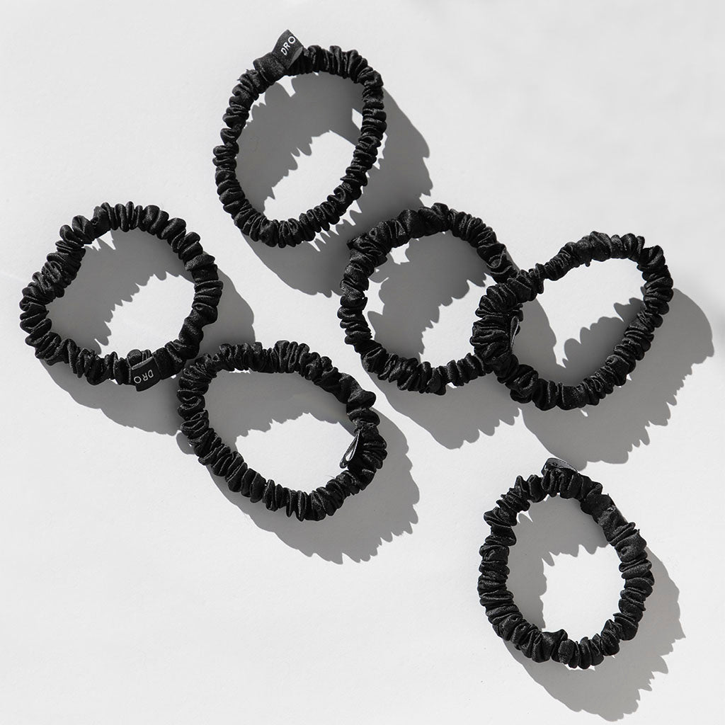 Six black Skinny Scrunchie Set - Black Jade hair ties by drowsysleepco are arranged on a light grey surface, each with a small black tag. Made from 22 momme silk, these elastic bands have a slightly ruffled texture and cast soft shadows.