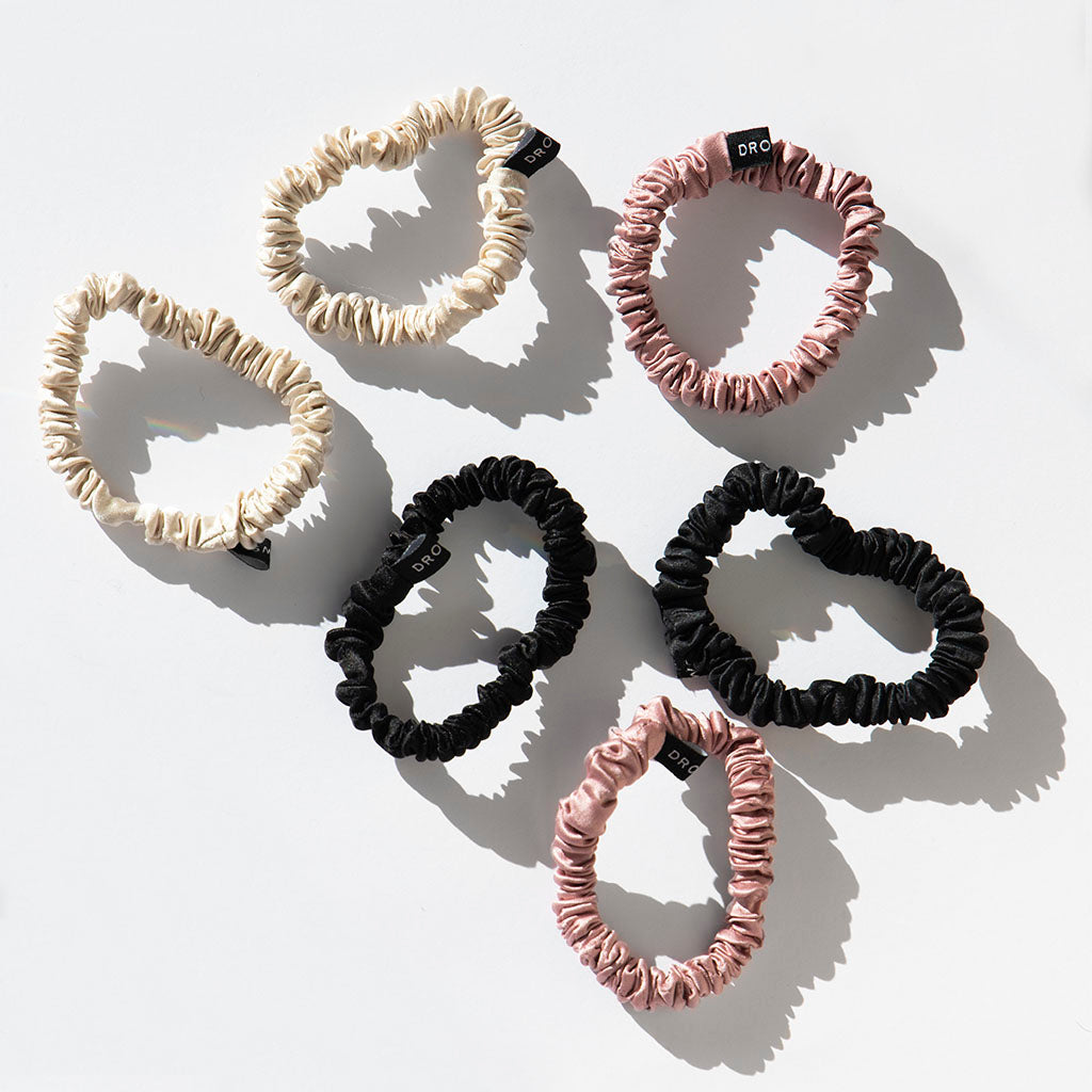 The drowsysleepco Skinny Scrunchie Set in Rose, Dusty, and Black includes six 22-momme silk scrunchies, each with a small black tag—perfect for hair health and arranged on a white surface with soft sunlight shadows.