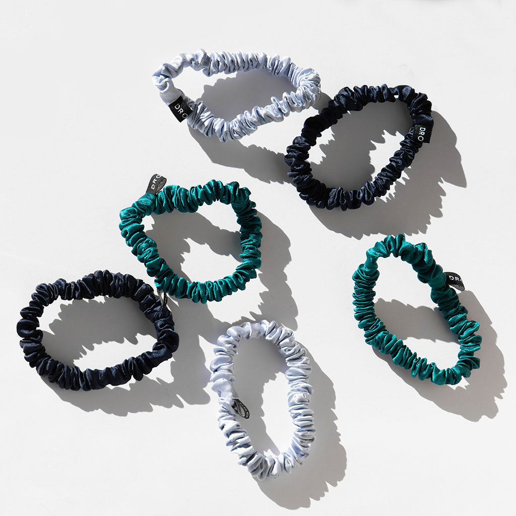 The drowsysleepco Skinny Scrunchie Set in Blue & Green Shades includes six gathered silk scrunchies—light blue, dark blue, and teal—each with a black tag, arranged on a white surface and ideal for hair health.