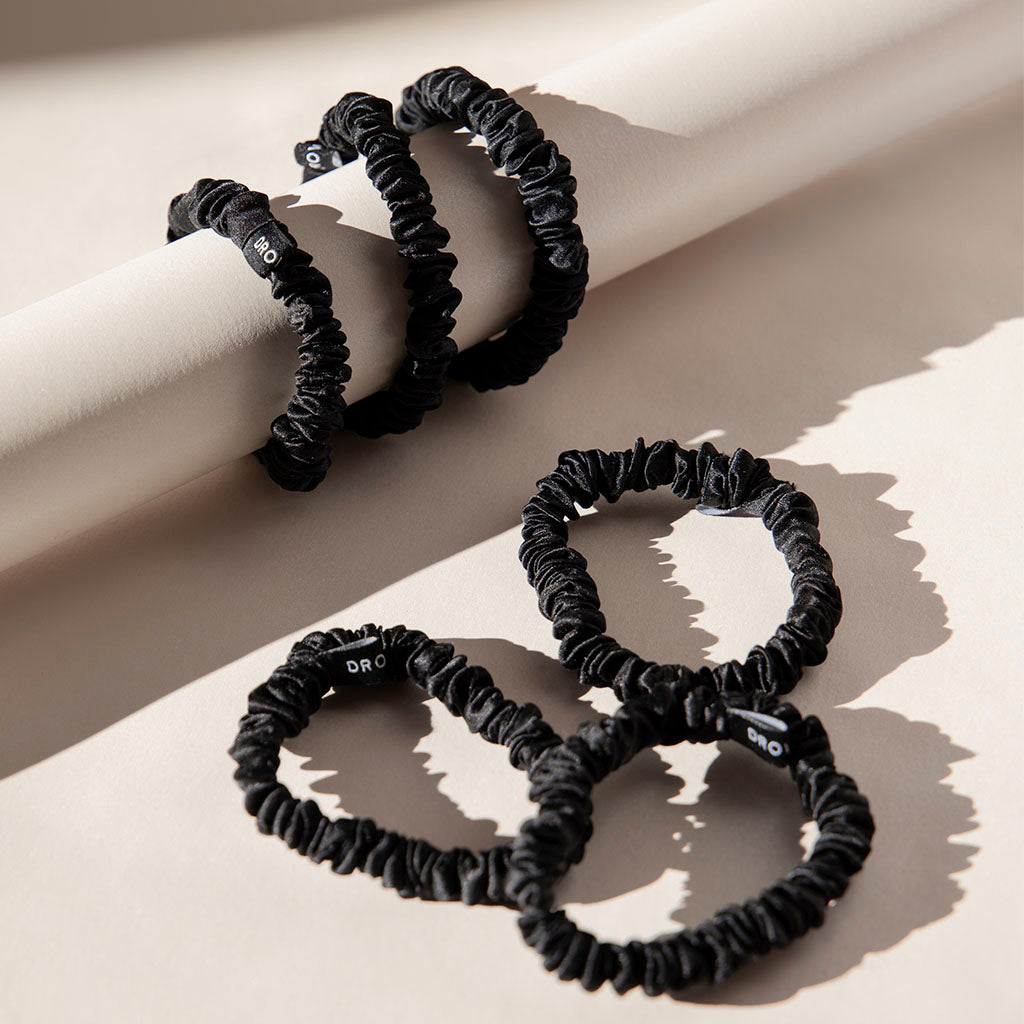 The drowsysleepco Skinny Scrunchie Set - Black Jade features four black, silk scrunchies wrapped around a beige rod, with three more arranged below on a flat surface, all in bright lighting with soft shadows.