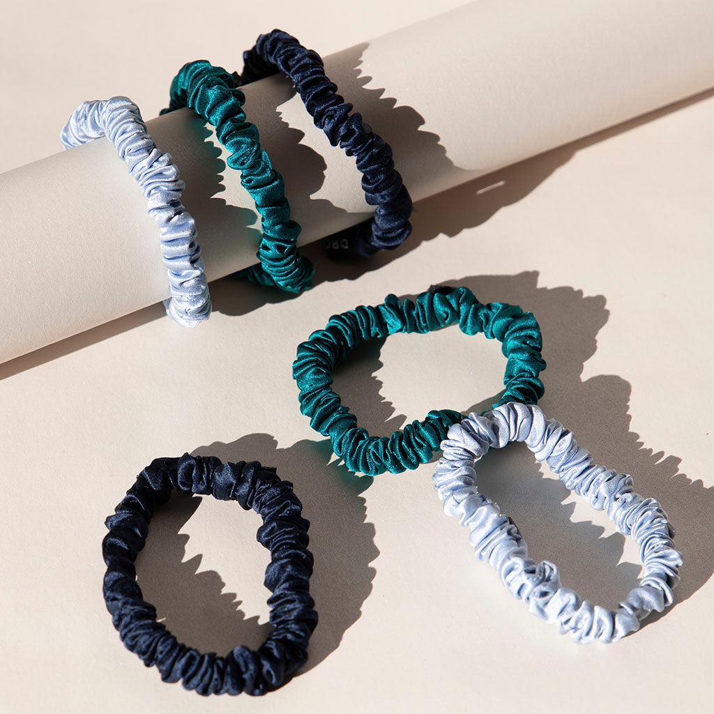 The drowsysleepco Skinny Scrunchie Set - Blue & Green Shades features five silk hair ties in navy, teal, and light blue, displayed on a cream surface—some draped on a cylinder, others flat—designed to support hair health.