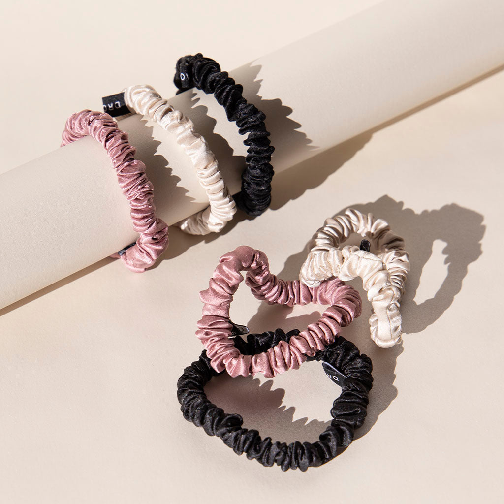 The drowsysleepco Skinny Scrunchie Set - Rose, Dusty, Black features three pink, two black, and two cream silk scrunchies (22 momme), artfully arranged on a cream cylinder with soft shadows—ideal for gentle hair care.