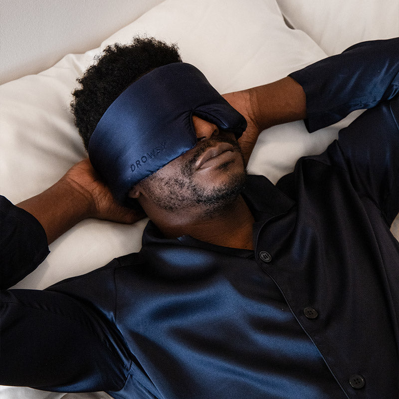 A man relaxes with his hands behind his head, wearing the drowsysleepco Sleep Week Ritual satin sleep mask and pajama top as he lies on a white silk pillowcase, looking peaceful and ready for sleep.