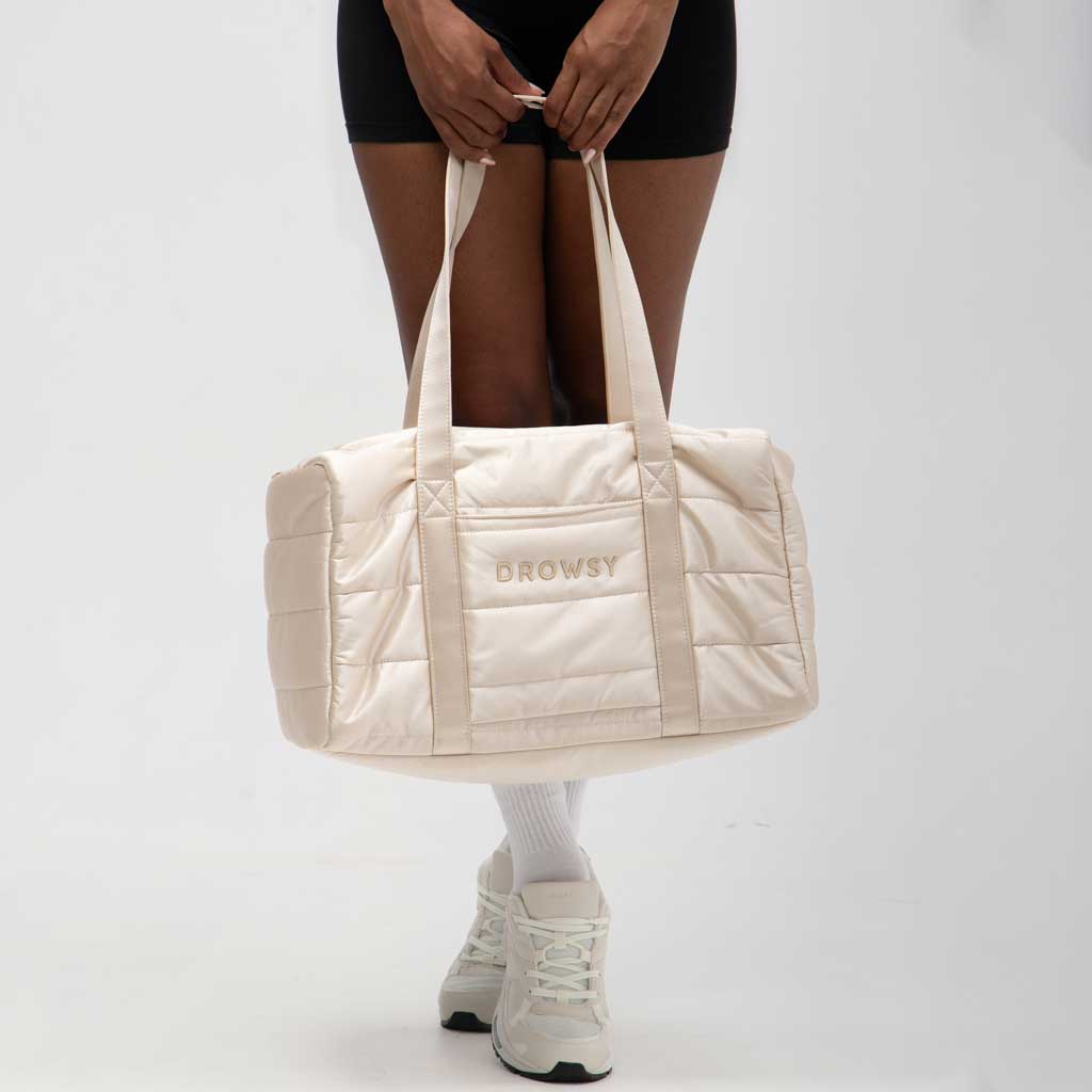 A person holds the drowsysleepco Perfect Escape Bundle - Dusty Gold, a large cream quilted duffel with DROWSY embroidered on the side—ideal for your silk sleep mask. They're in black shorts and white sneakers against a plain white background.