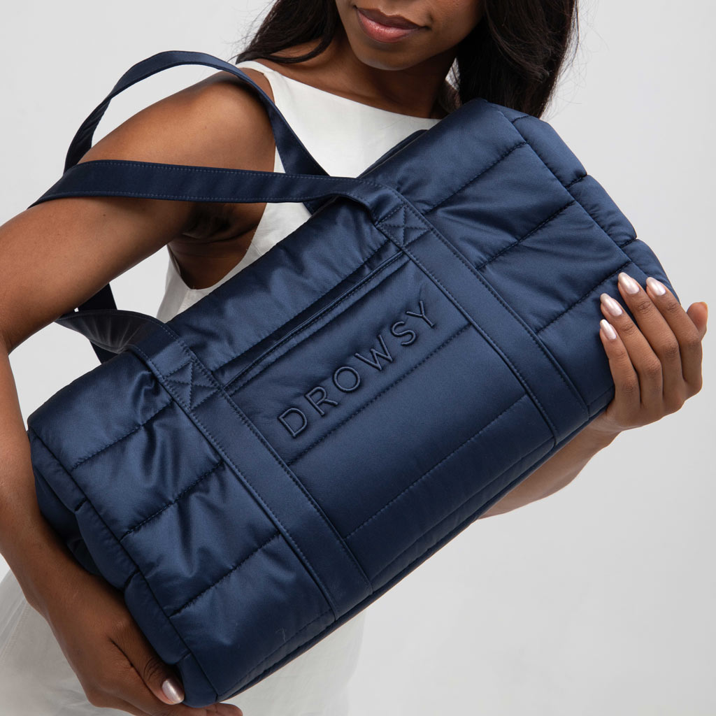 A woman in a sleeveless white top holds the drowsysleepco Perfect Escape Bundle - Midnight Blue duffel—ideal for carrying your silk travel essentials and Silk Sleep Mask for beauty sleep wherever you go.