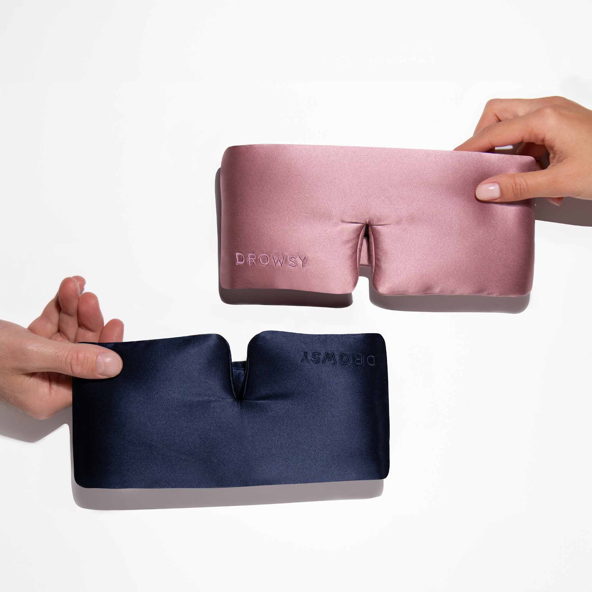 Two hands each hold a drowsysleepco Lovers Pack luxury silk sleep mask—one pink, one navy blue—against a white background.