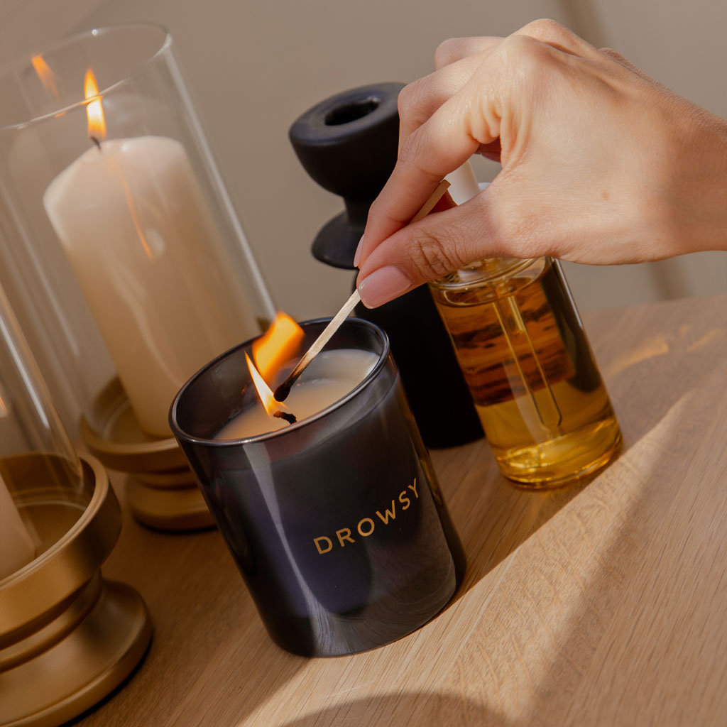 A hand lights the drowsysleepco Sleep Ritual Candle 220g, made with natural essential oils, using a match. The black candle sits on a wooden surface beside a glass oil bottle and other lit candles in decorative holders.