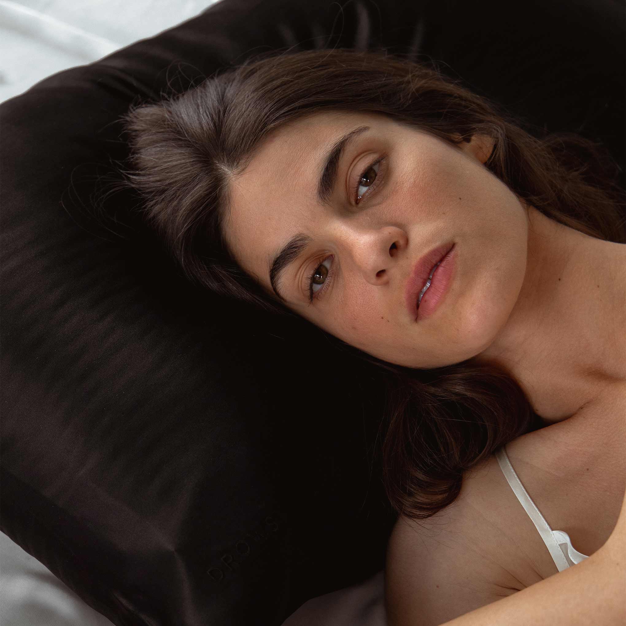 A woman with long dark hair lies on a black pillow, looking calmly at the camera. She is wearing a white strap top and appears relaxed, with soft natural light illuminating her face.