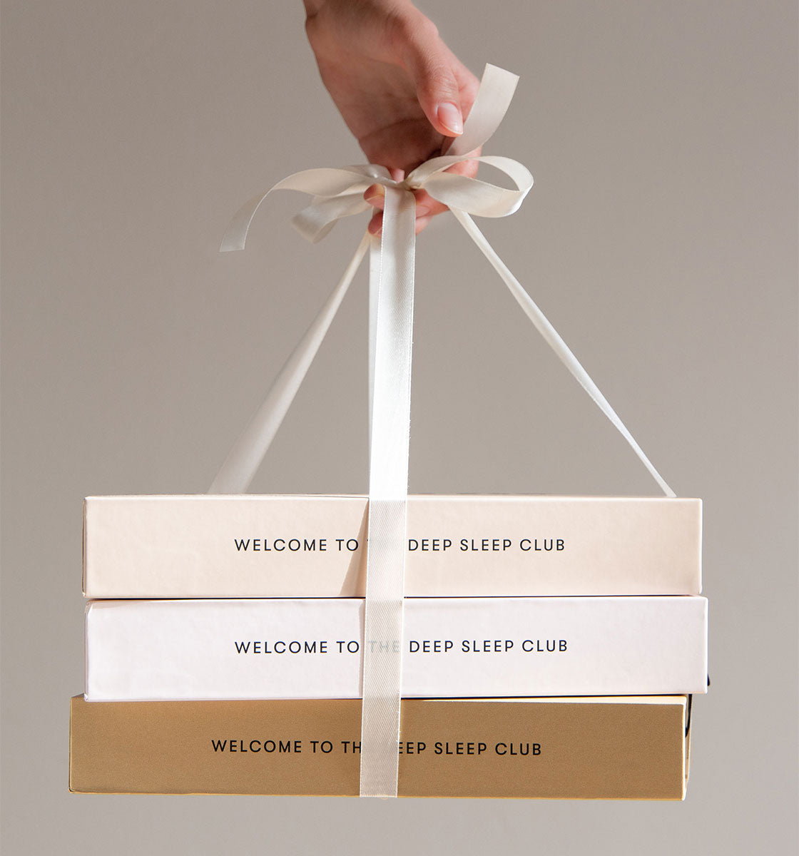 A hand holds three stacked boxes tied with a white ribbon. The boxes are cream, white, and gold, each labeled WELCOME TO THE DEEP SLEEP CLUB.