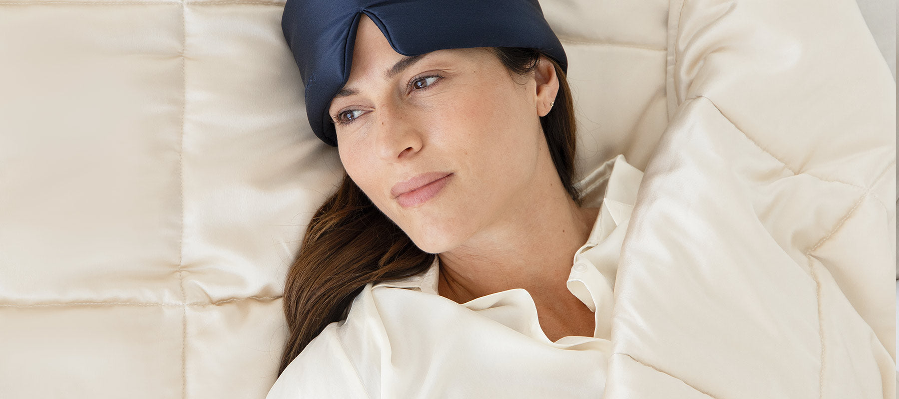 A woman wearing a navy blue sleep mask on her forehead lies in bed under a cream-colored comforter, looking relaxed and thoughtful.