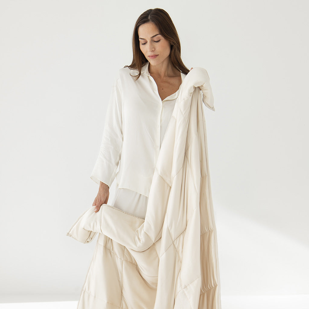 A woman in a loose white shirt holds the drowsysleepco Dusty Gold Silk Weighted Blanket, draped over her arm, while looking down gently against a plain white background.