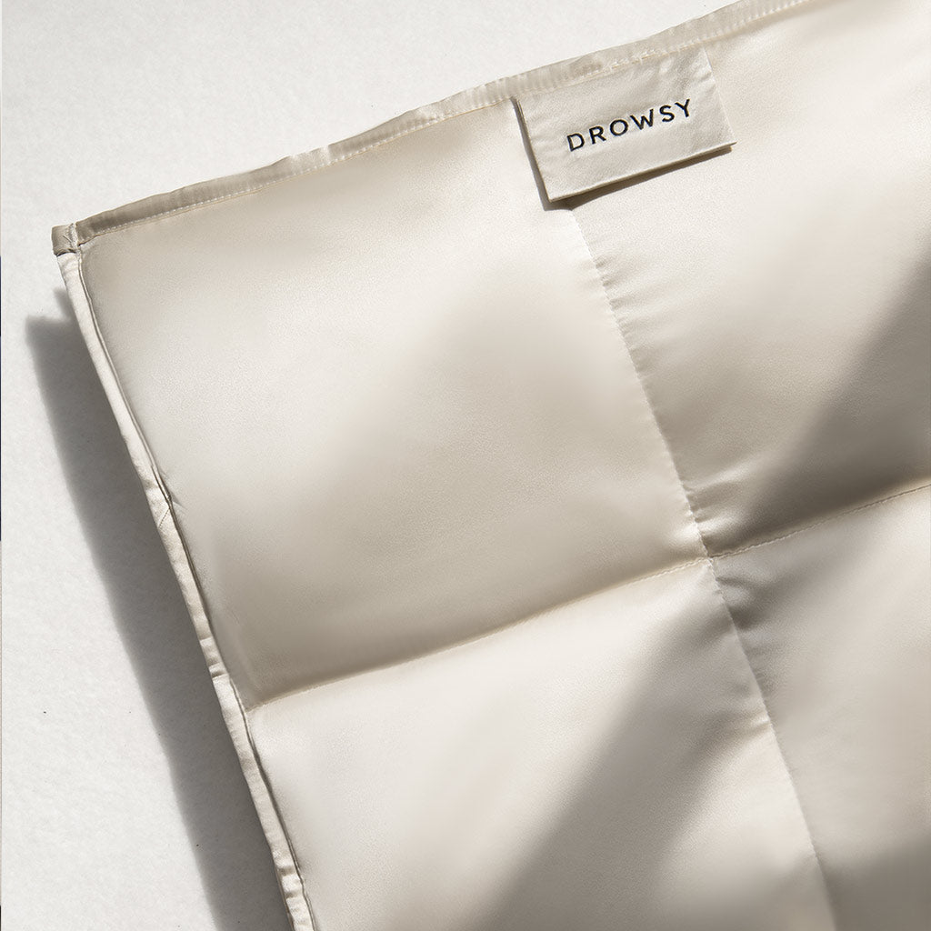 A close-up of the Dusty Gold Silk Weighted Blanket by drowsysleepco, featuring a quilted, cream-colored mulberry silk design and a DROWSY fabric tag on the upper right corner, resting on a soft white surface.