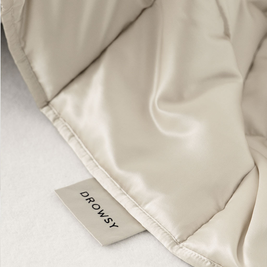 A close-up of the Dusty Gold Silk Weighted Blanket from drowsysleepco, showing its smooth, silky beige fabric and a tag reading DROWSY, displayed on a white surface.