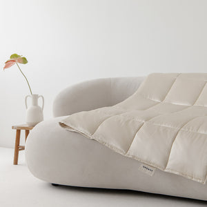The drowsysleepco Dusty Gold Silk Weighted Blanket is draped over a modern curved white sofa, with a small wooden table in the background holding a white vase and a single green-pink leaf, creating a minimal, bright aesthetic.