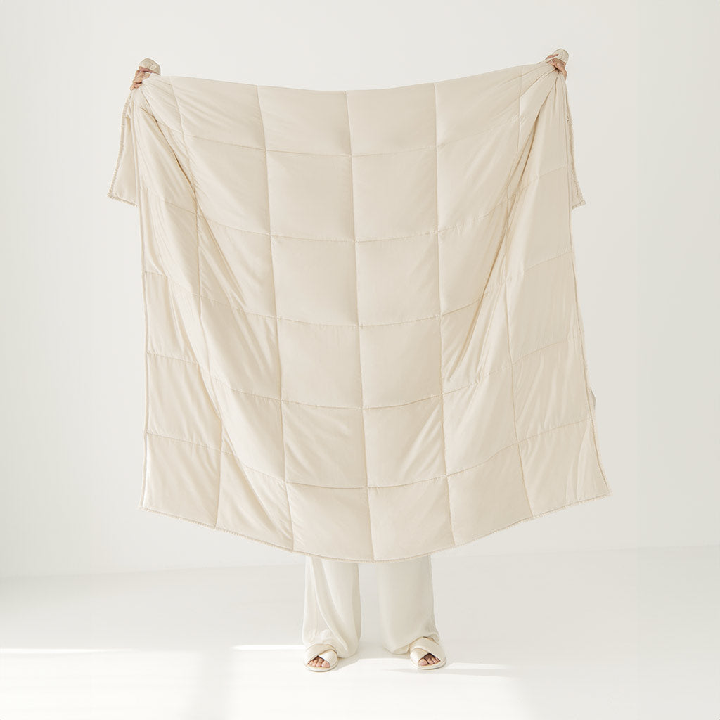 A person holds up the drowsysleepco Dusty Gold Silk Weighted Blanket, covering most of their body with only their hands, feet, and part of their legs visible against a plain white background.