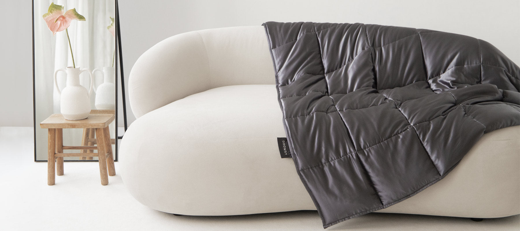 A modern, white curved sofa with a dark gray quilt draped over one side sits beside a wooden stool holding two white vases and a pink flower, with a large mirror in the background.