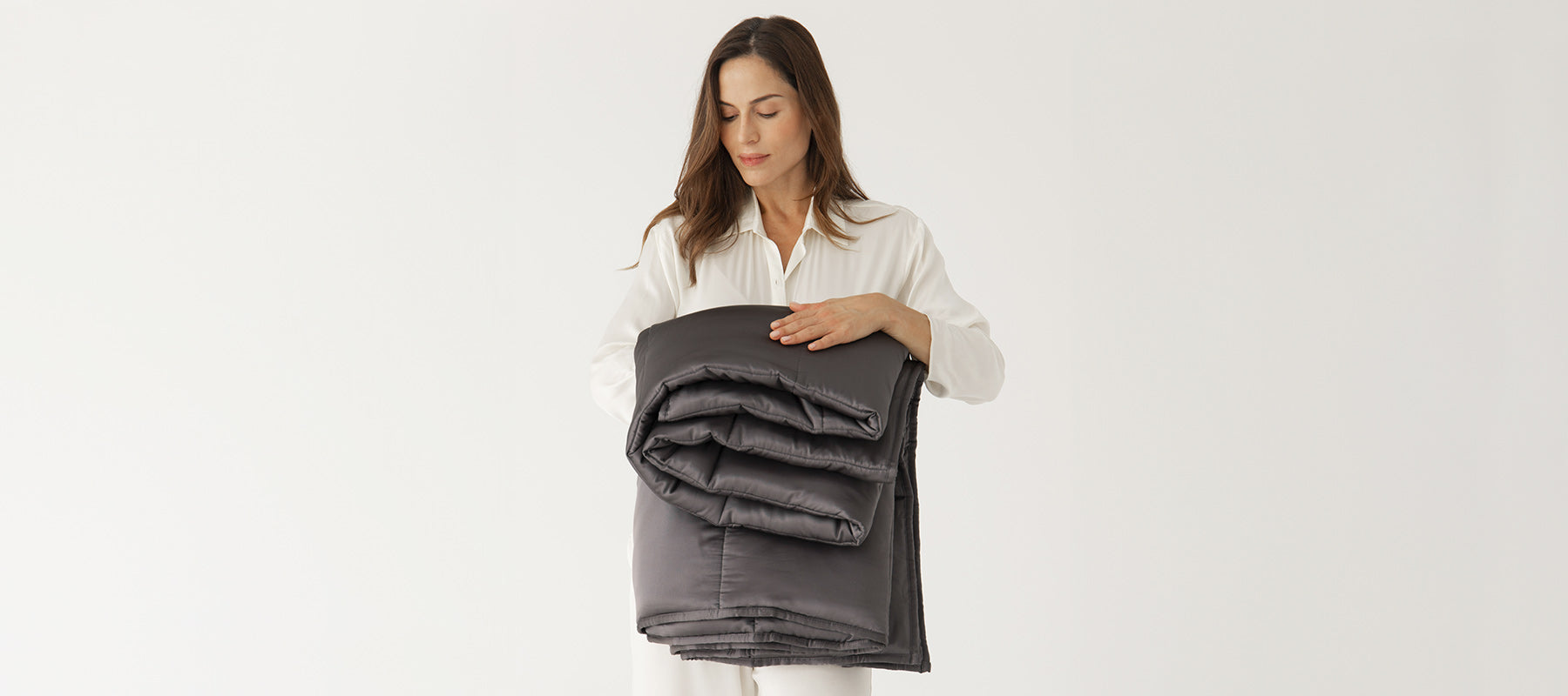 A woman in white pajamas stands against a plain white background, holding a neatly folded dark gray weighted blanket in her arms and looking down at it with a gentle expression.