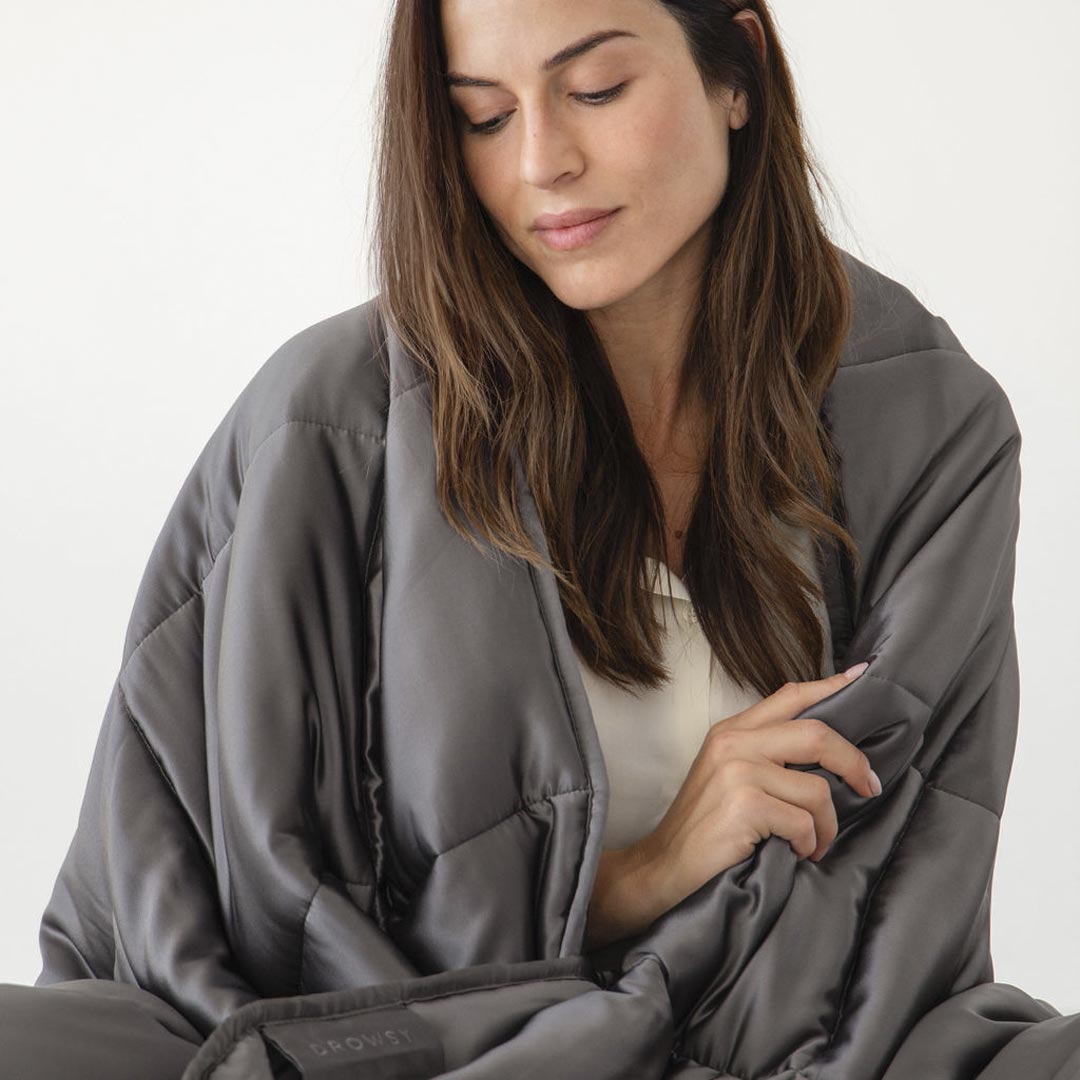 A woman with long brown hair sits wrapped in the drowsysleepco Moonlight Shadow Silk Weighted Blanket, looking down gently against a plain light background.