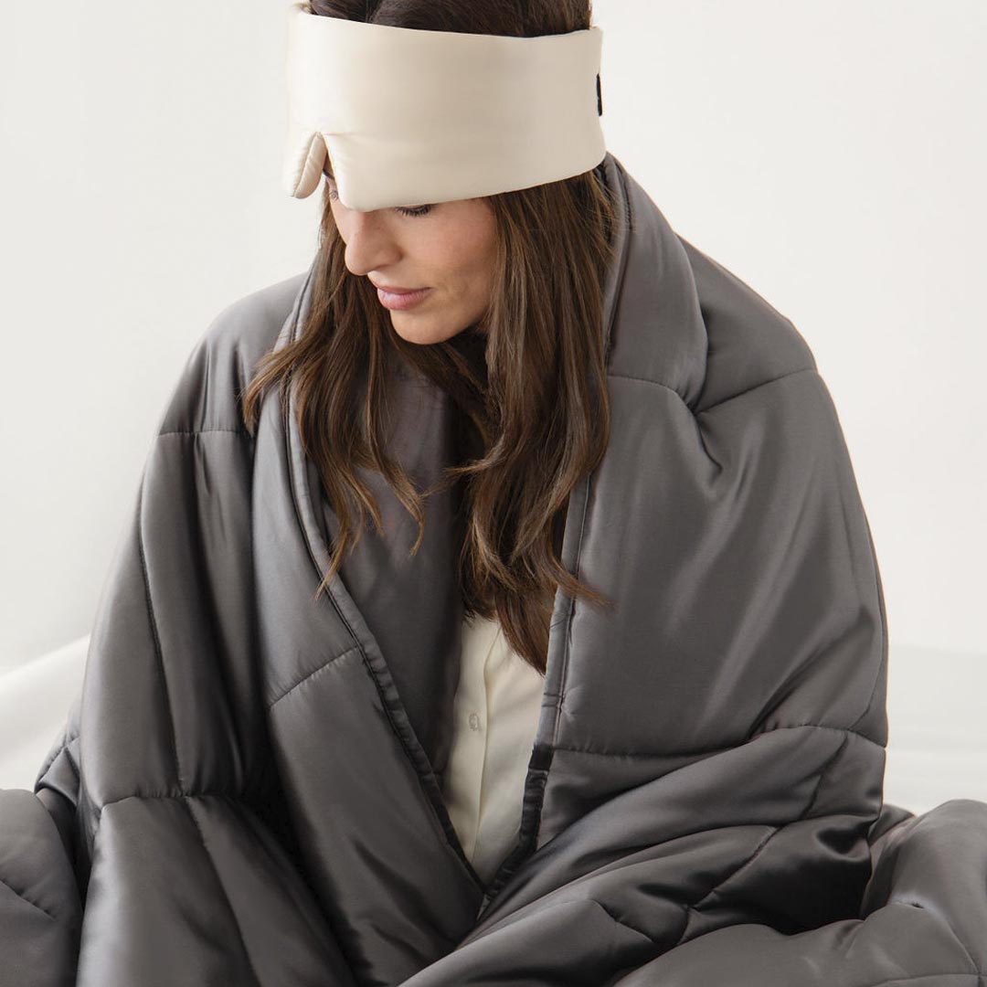 A woman sits wrapped in the drowsysleepco Moonlight Shadow Silk Weighted Blanket, wearing a cream sleep mask. Her long brown hair peeks out as she looks down, set against a plain, light background.