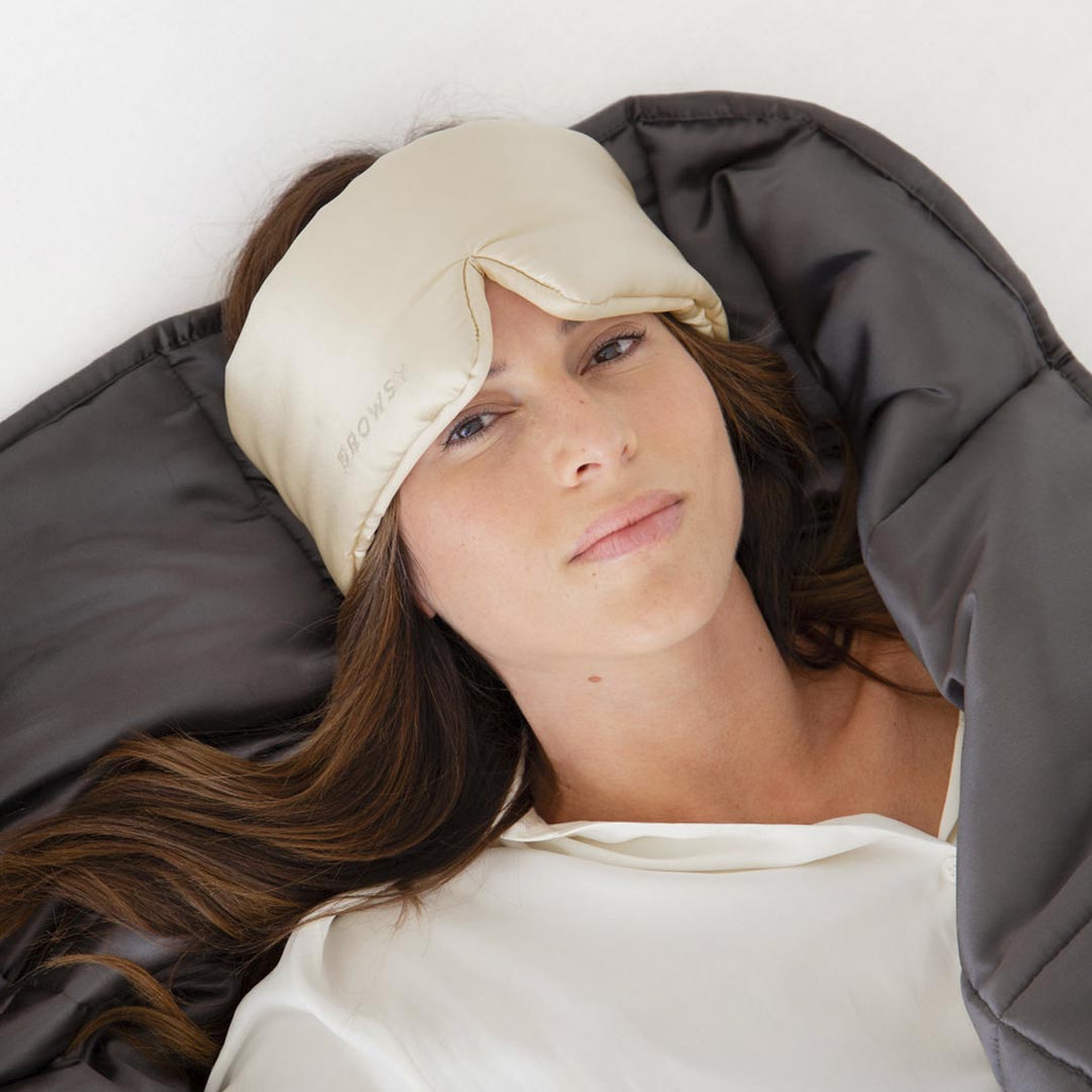 A woman with long brown hair lies on her back wearing a beige sleep mask and a white top, partially covered by a dark gray blanket. She looks relaxed and restful.