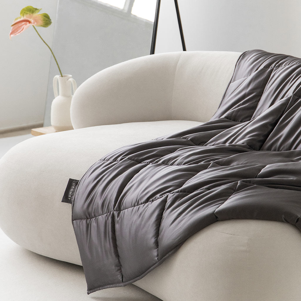 The drowsysleepco Moonlight Shadow Silk Weighted Blanket in dark gray is draped over a modern cream curved sofa in a minimalistic room, with a vase holding a single flower in the background.