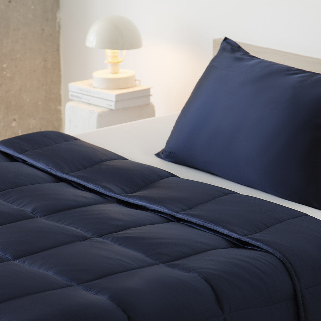 A neatly made bed with the drowsysleepco Midnight Blue Silk Weighted Blanket and pillow, beside a white nightstand with books and a round white lamp, creates an inviting space for deep relaxation in a modern minimalist bedroom.