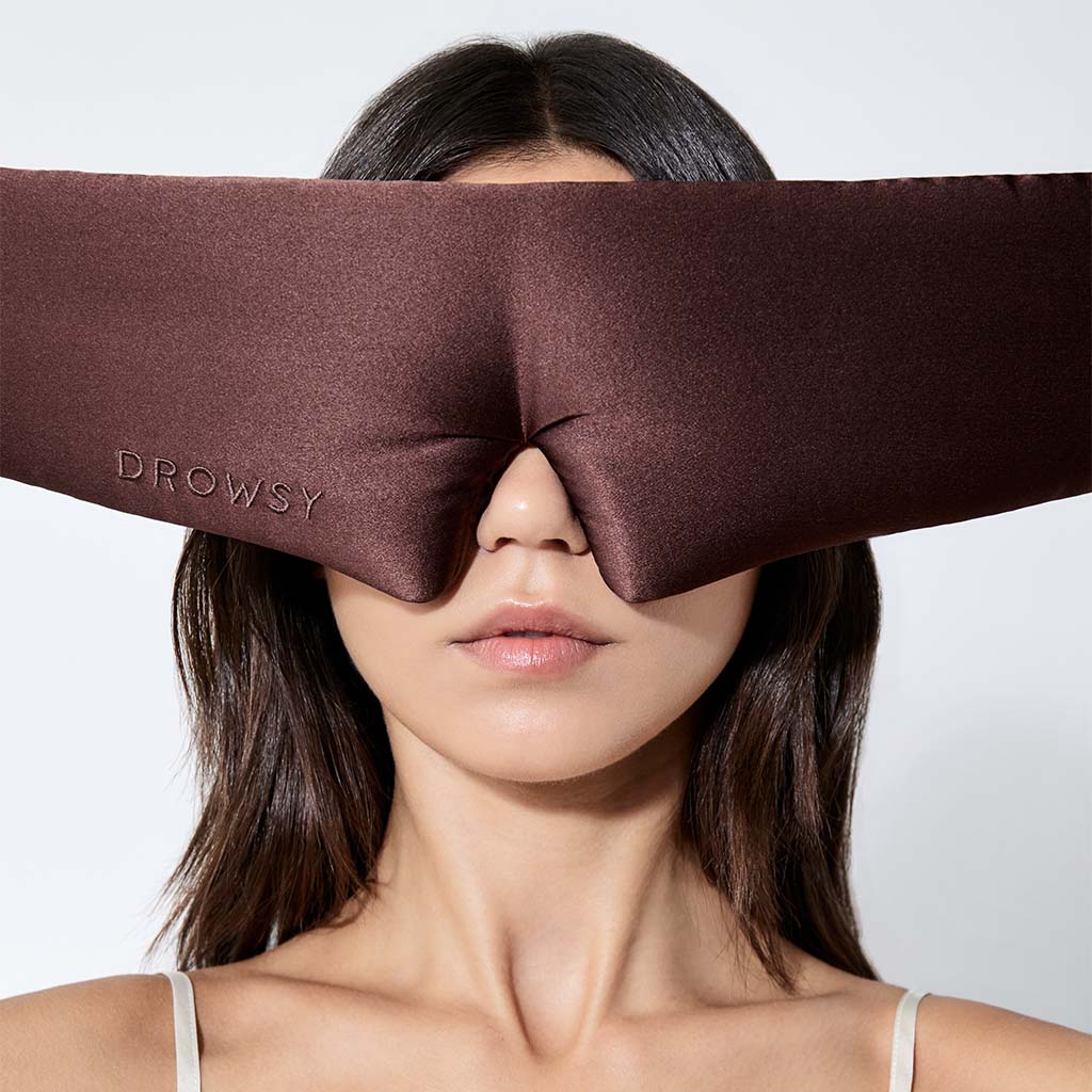 A woman with straight dark hair faces forward against a white background, wearing the large brown Rajah Sleep Mask by drowsysleepco. "DROWSY" is embroidered on the silk mask, which covers her eyes and nose.