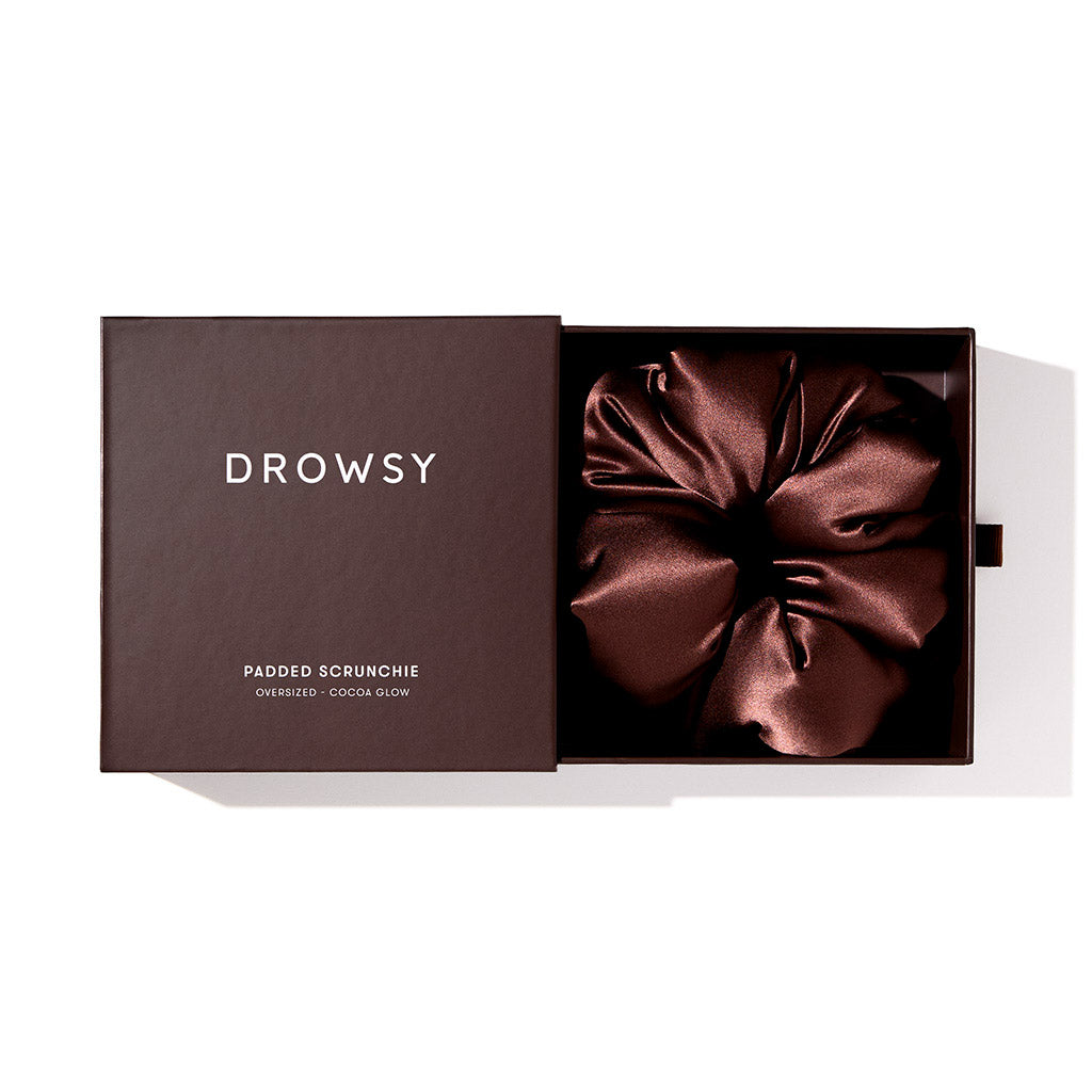 A Cocoa Glow - Padded Scrunchie by drowsysleepco rests in an open box labeled with its name. This oversized, dark brown silk hair accessory is displayed on a white background—ideal for protecting hair during sleep.