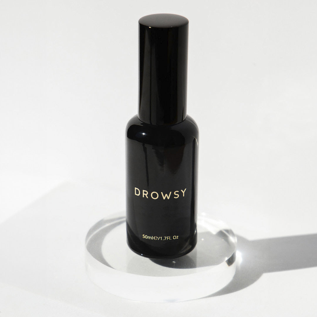 A black bottle labeled DROWSY sits on a clear, round platform against a white background. The bottle is upright, sleek, and minimalist with gold text and a shiny finish.
