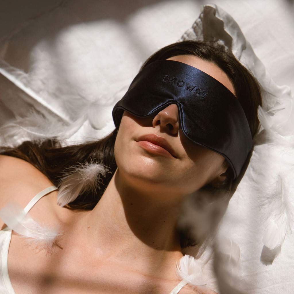 A woman lies on a bed wearing a black sleep mask with the word Drowsy on it. White feathers are scattered around her, and soft light filters through, creating a calm, restful atmosphere.