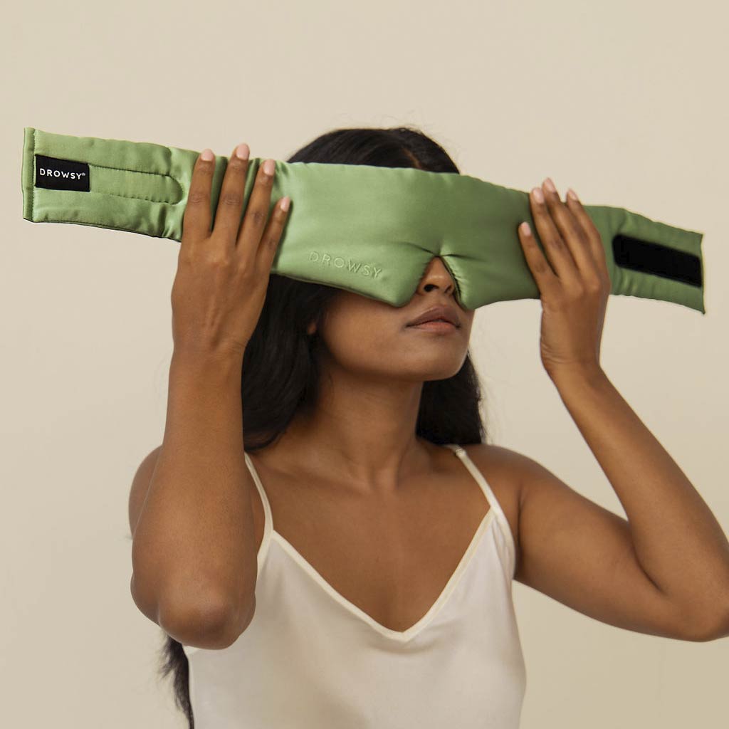 A woman in a white camisole holds a Pistachio Green silk sleep mask from drowsysleepco over her eyes, enhancing her beauty sleep against a plain beige background.