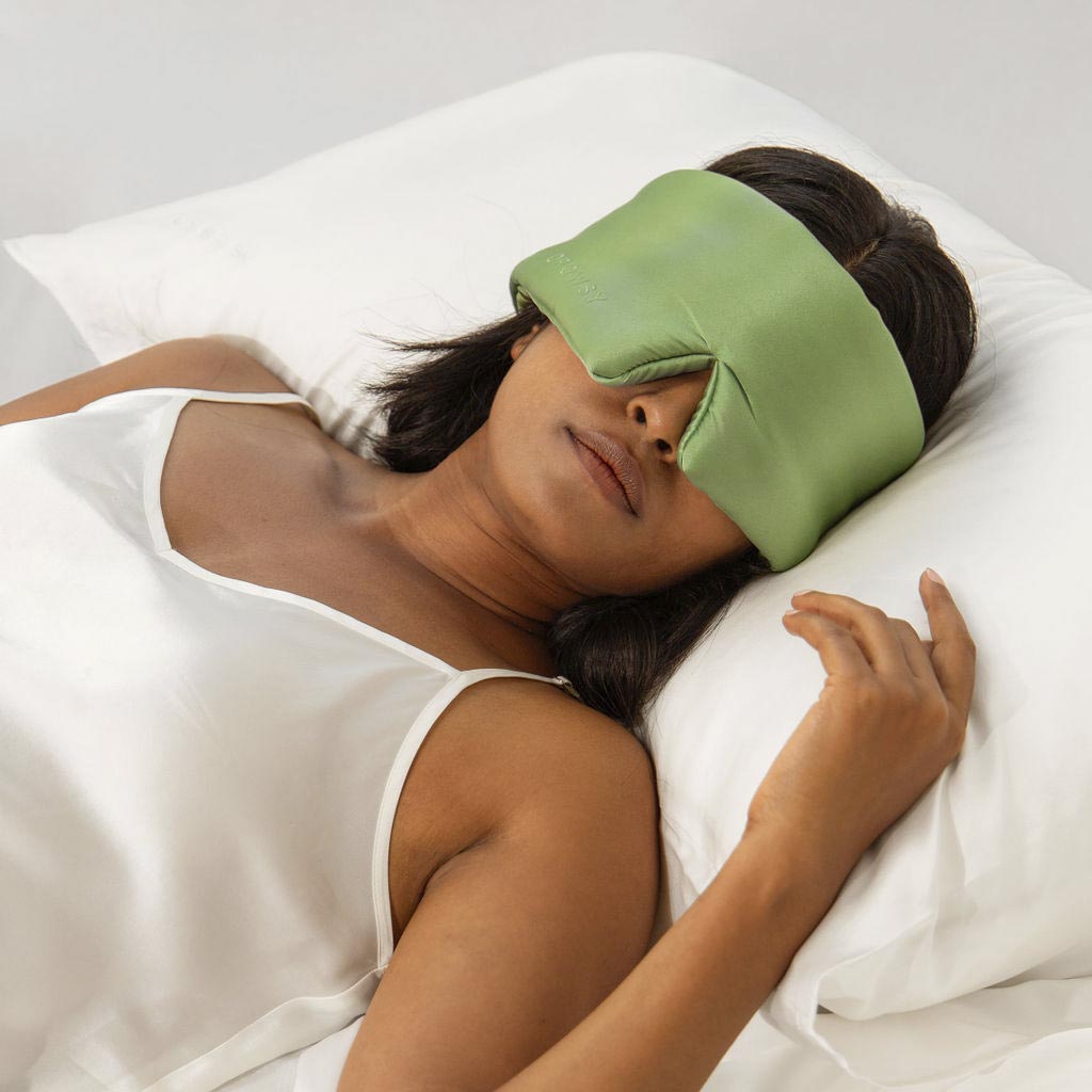 A woman in a white satin camisole lies on a white pillow, wearing a large green sleep mask that covers her eyes and part of her nose. She appears relaxed and is resting on her back.
